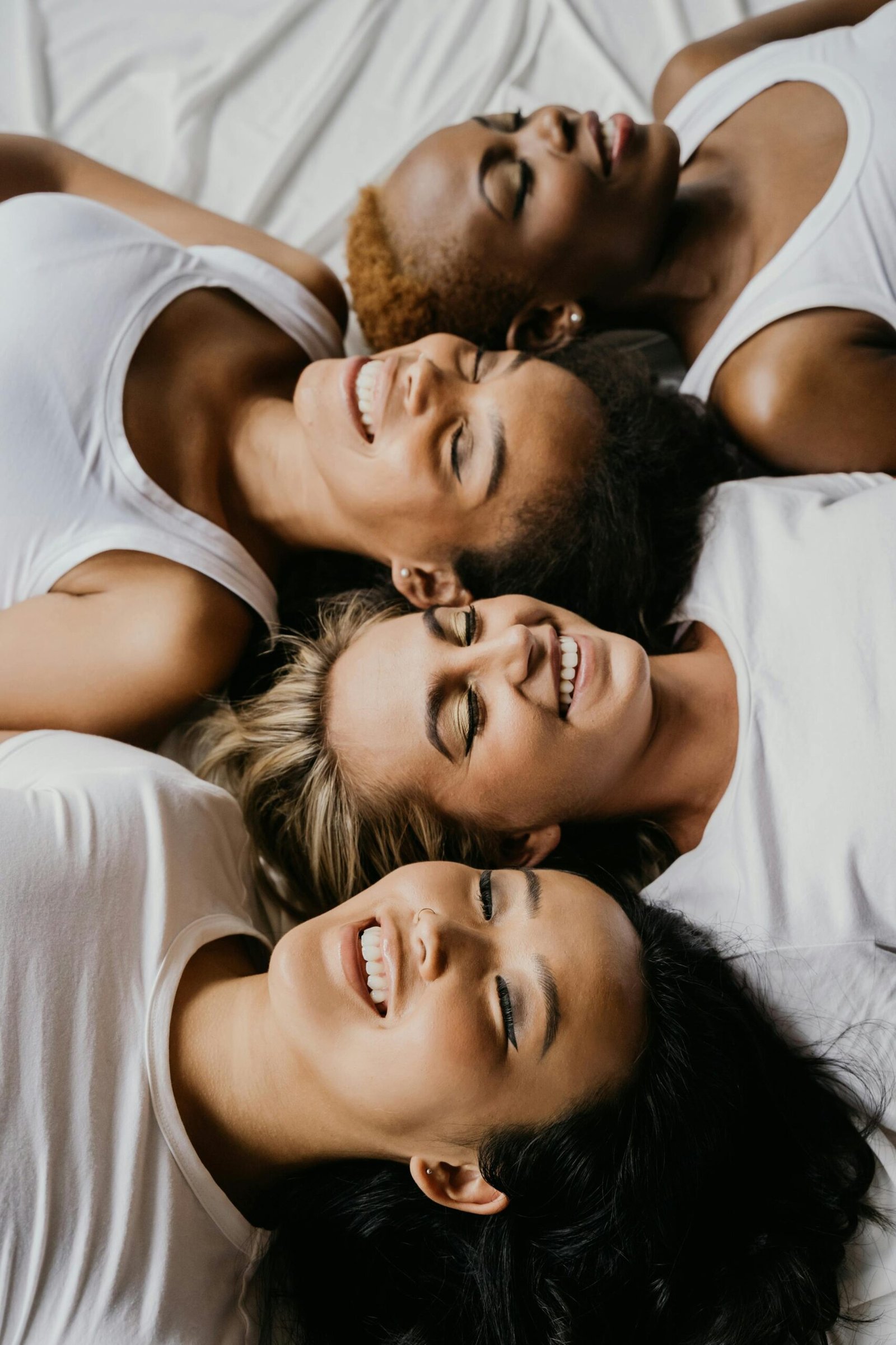 Four diverse women lying on a bed, smiling together, representing unity and friendship.