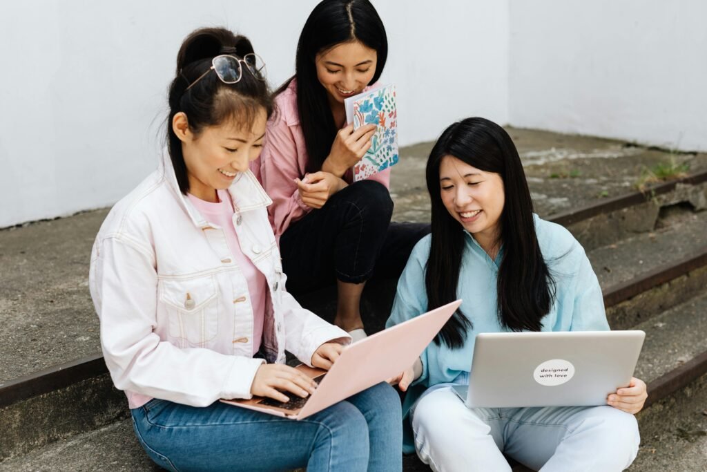 Three smiling women sitting on outdoor steps, working together with laptops and a journal.